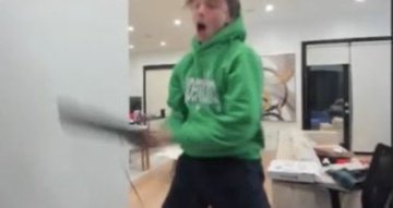 A man in a green hoodie is seen swinging a laptop in a fit of rage, likely reacting to a crypto market crash. The blurry motion of the laptop conveys intense frustration, fitting the "biggest crash outs" theme tied to cryptocurrency losses.