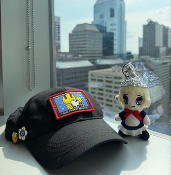 The image features a black baseball cap with a cartoon character design, placed next to a small stuffed animal doll wearing a dress and hat. The caption of the image reads "Don't worry, I don't know why you're here".