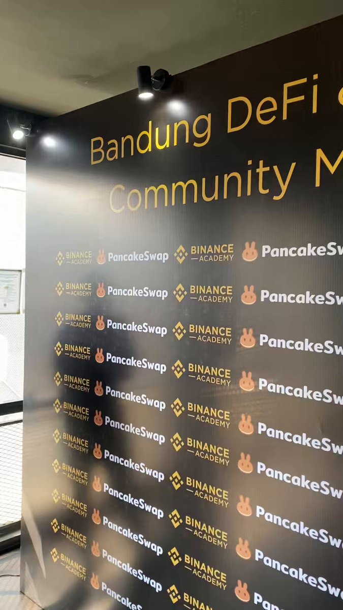 The image shows a large black wall with white text, advertising various logos for different companies such as Bitcoin, PancakeSwap, and BinanceAcademy. The text reads "Joining the crowd and community @BinanceAcademy x #PancakeSwap massive opportunity & new friends". This suggests that there is an event or gathering related to cryptocurrency and blockchain technology taking place at this location.