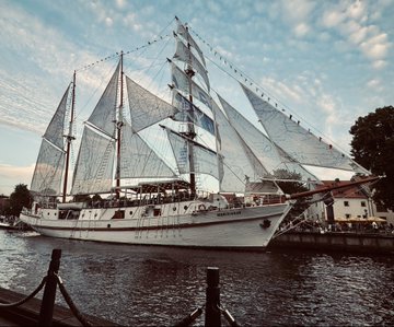 A large white sailboat with multiple masts docked in calm water during what appears to be sunset.