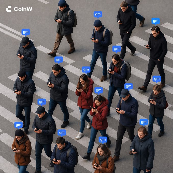 An aerial view shows a busy crosswalk filled with pedestrians in winter clothing, nearly all absorbed in their smartphones, with blue "gm" speech bubbles floating above each person. The CoinW logo appears in the top-left corner, tying the everyday scene of people checking their phones to the crypto community's popular "gm" greeting.