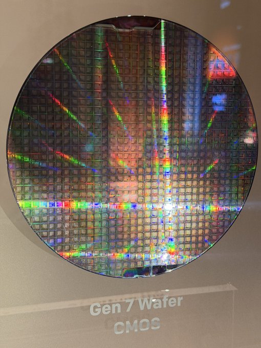 A close-up photograph of a circular silicon wafer with iridescent rainbow reflections on its grid-like surface, displayed behind glass with the text 'Gen 7 Wafer CMOS' visible on the label.