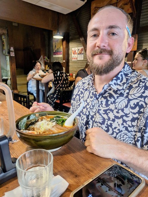 A smiling bearded man in a blue and white floral shirt sits at a wooden counter enjoying a large bowl of ramen with chopsticks and a spoon, while a glass of water and smartphone rest nearby in what appears to be a cozy Japanese restaurant with other diners visible in the background. The image perfectly captures the birthday reflection in the caption—nostalgically celebrating turning 31 by reminiscing about a past ramen shop experience in Japan.