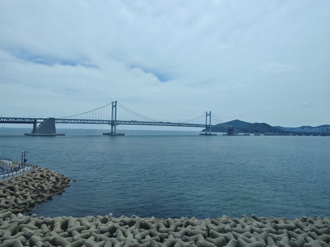 A long suspension bridge spans across the calm sea under a cloudy sky, with concrete tetrapods lining the rocky shore in the foreground, capturing a scenic Korean coastal view that complements the post's celebration of Liberation Day in Korea.