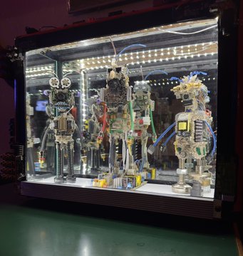 An illuminated display case containing four robotic figures constructed from electronic components and mechanical parts, with blue tubing and wires visible.