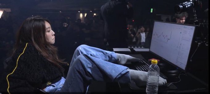 A young woman sits with her feet up on a desk in a crowded gaming or esports event, looking at a monitor displaying a price graph, dressed in a black jacket and jeans.