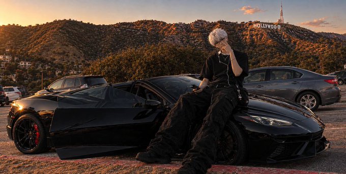 A person with silver hair wearing all black leans against a black Chevrolet Corvette parked at an overlook, with the iconic Hollywood sign visible on the hillside in the background at sunset.