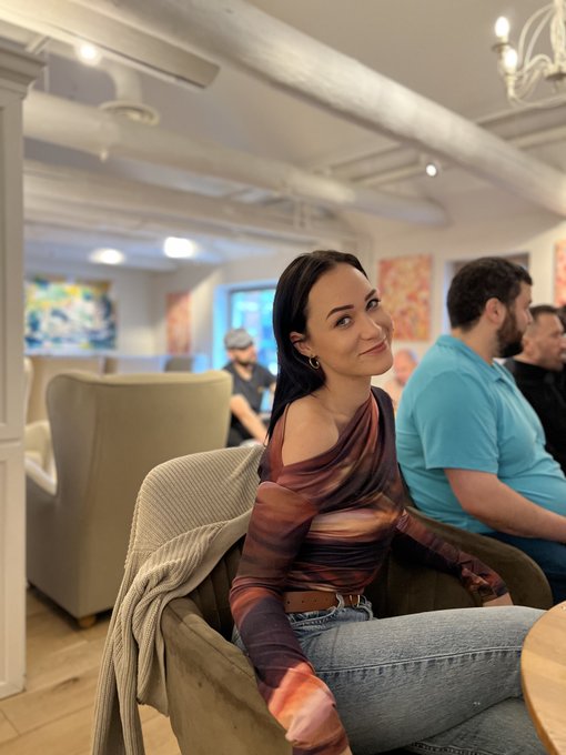 A woman with dark hair in a sheer multicolored long-sleeve top and jeans smiles at the camera while sitting in an armchair, with a man in a blue polo shirt seated behind her in a bright interior space.