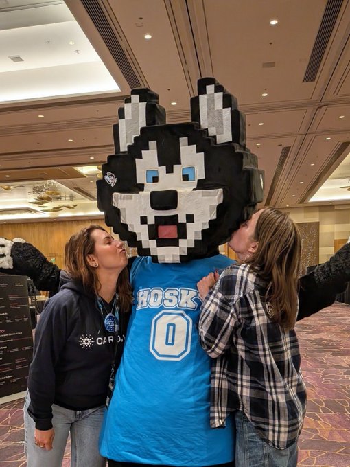 Two women kissing a mascot dressed as a Husky wearing a blue jersey with the number 0 in what appears to be a hotel or conference setting.