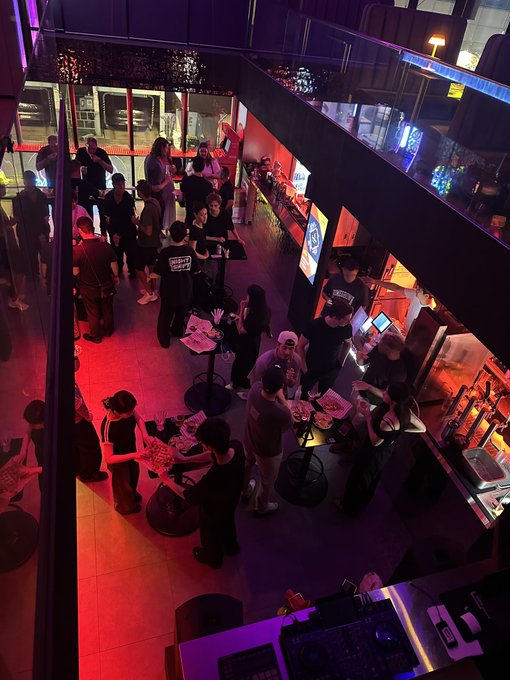 An overhead view of a dimly lit bar or club with red and purple lighting, where patrons gather around tables and at the bar.