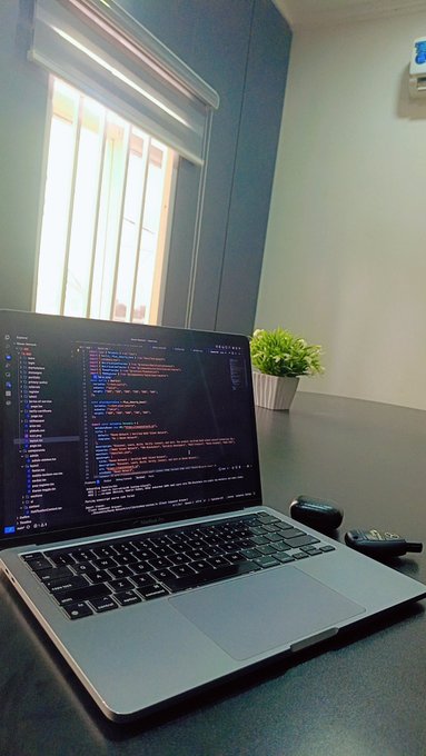 A MacBook displaying colorful code in an editor sits on a desk near a window with a small potted plant and wireless earbuds nearby—capturing the "Gm" (good morning) vibe of starting a productive coding session.