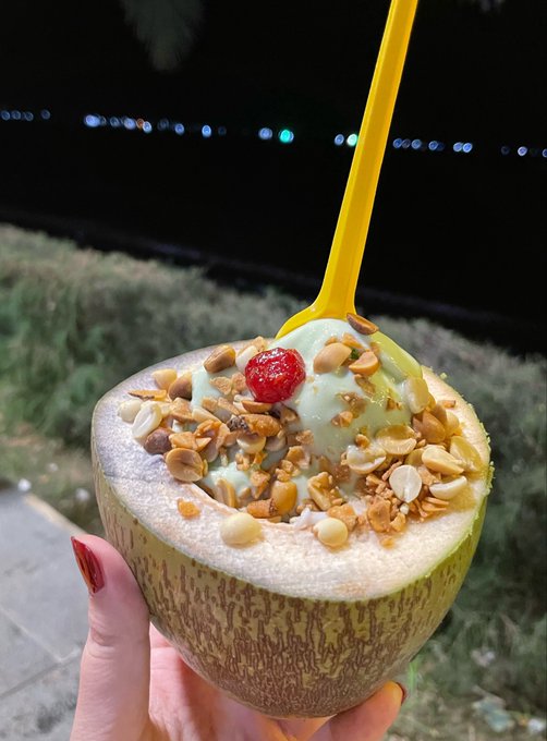 A hand holding a halved cantaloupe filled with a creamy dessert topped with nuts and a strawberry, with a yellow spoon sticking out, photographed at night.