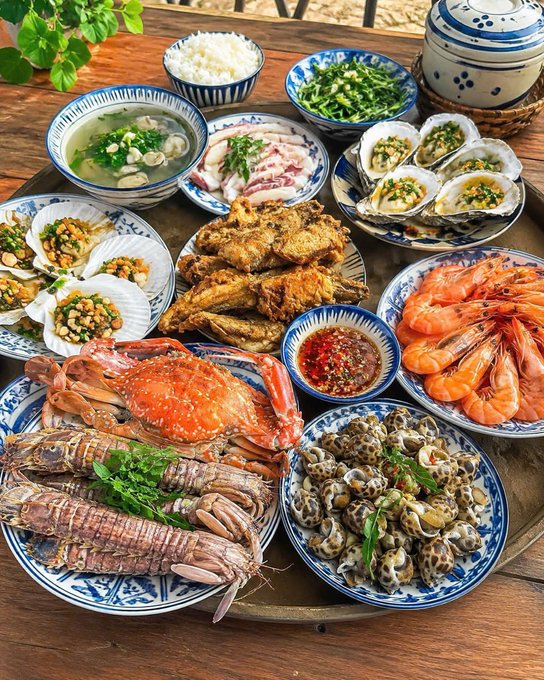 A large spread of Vietnamese seafood dishes is arranged on a wooden table, including crab, shrimp, mantis shrimp, snails, oysters, scallops, fried fish, and rice.