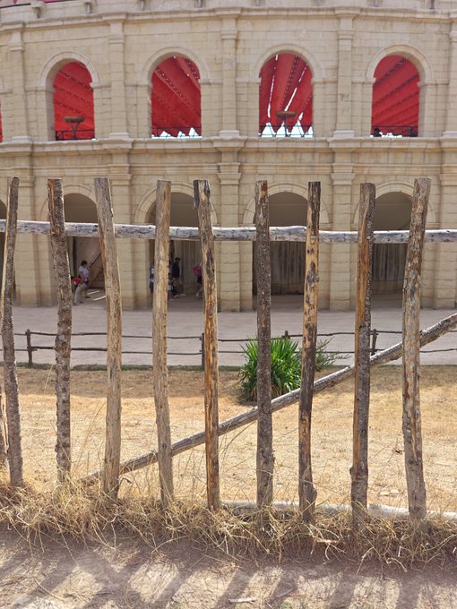 A wooden fence made of vertical posts in front of a historic building with arched openings containing bright red curtains or fabric, with a few people visible in the background courtyard.
