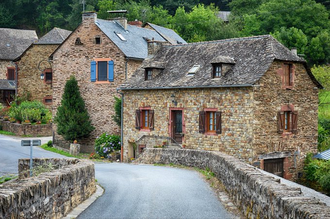 Charming stone houses with wooden shutters line a quiet road in a rural European village, with one prominent building featuring blue shutters and surrounded by greenery.