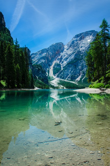 A scenic landscape photograph of a turquoise alpine lake reflecting snow-capped mountains and evergreen trees under a blue sky.