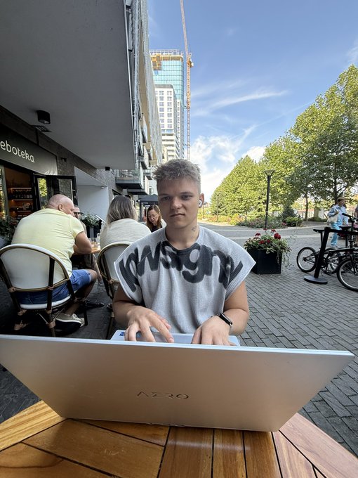 A young man in a "Wrangler" sweatshirt sits at an outdoor café working on an AZIO laptop, styled as a relatable "gm" (good morning) moment for the web3 community. The "Gm web3 ex" caption ties the casual, on-the-go crypto lifestyle aesthetic to an everyday remote work vibe.