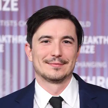 A man in a dark blue suit and black tie stands smiling in front of a backdrop with partial text including "AKTHRO" and "PRIZE," likely at an awards or promotional event. The image does not directly relate to the accompanying post about Robinhood Chain's 24-hour DEX volume surpassing $500 million, as no Robinhood branding, financial figures, or cryptocurrency-related content is visible in the photo.