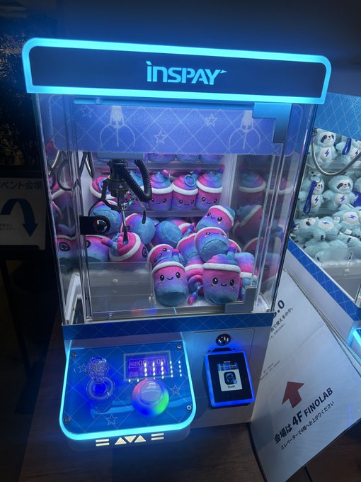 Illuminated 'INSPAY' claw machine filled with pink and blue cartoon plush toys, featuring a payment terminal.
