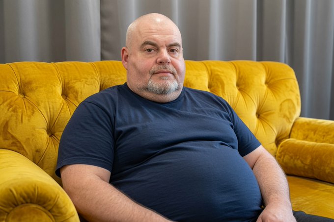 A bald, heavyset man with a gray goatee lounges lazily on a mustard-yellow tufted velvet sofa, wearing a dark navy t-shirt and looking off to the side with a relaxed, almost defiant expression. His sedentary pose perfectly embodies the post's tongue-in-cheek declaration of boycotting workouts until BSV is re-listed on "bucket shop" exchanges.