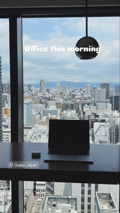 A view from an office desk in Osaka, Japan showing a laptop on the table with a city skyline through the window, captioned "Office this morning."