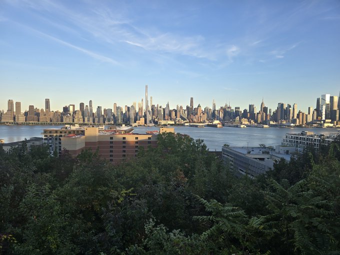 A daytime view of the Manhattan skyline, including tall skyscrapers, with a body of water in the midground and green trees and a brick building in the foreground.