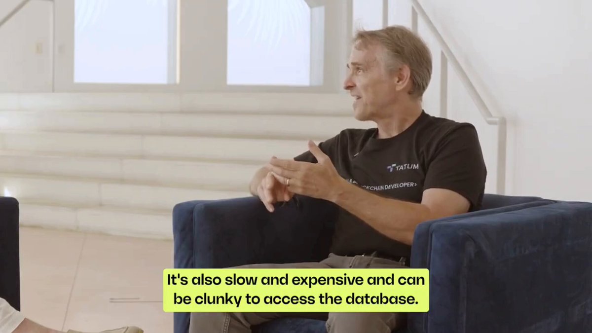 The image shows a man sitting in a chair with his hands on his lap while wearing a black shirt. The background of the image features white walls, which contrasts with the blue chair that the man is seated in. There are also two other chairs visible in the scene. The text "It's also slow and expensive to access the database" is written below the image, likely providing context or information about the content being displayed.