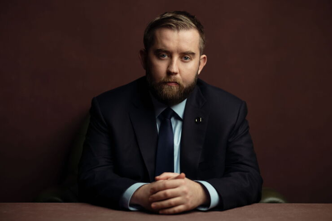 A bearded man in a dark suit and blue tie sits at a table with his hands clasped, looking directly at the camera against a dark maroon backdrop.