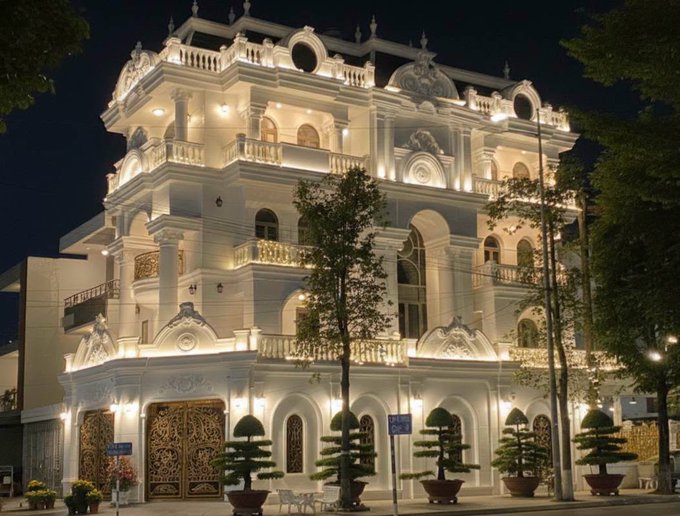 An ornate, multi-story white building illuminated with warm lights at night, featuring arched windows, balconies, and decorative landscaping in front.