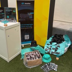 An exhibition display on artificial turf features a metal cabinet, a yellow and black 'NovaPlus' banner, scattered clothing, a hat, and various packaged items including chocolate snacks.