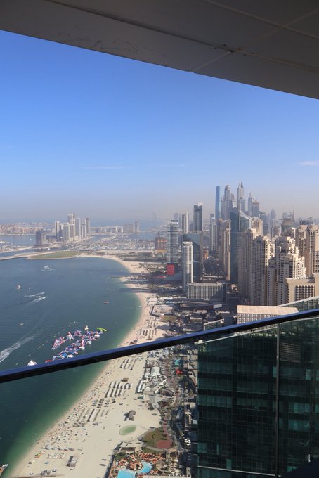 A view from inside the Burj Khalifa's observation deck in Dubai, UAE, looking down at the city skyline, beachfront, and boats along the coastline under a clear blue sky.