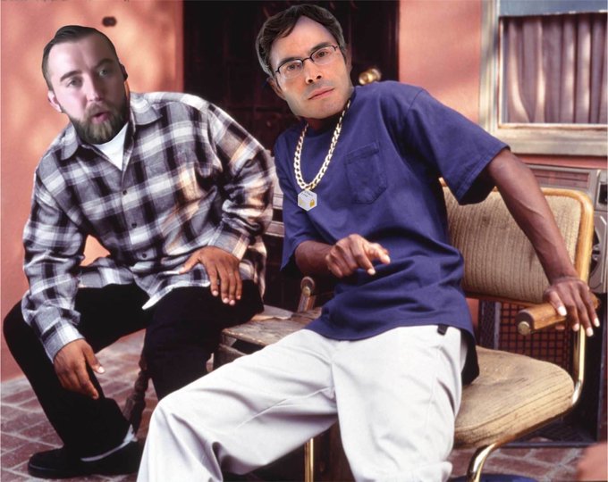 Two men are seated together in an outdoor setting, one wearing a plaid shirt and the other wearing glasses, a large gold chain, and a dark blue shirt.