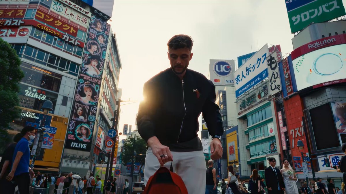 A man in a black Yoda jacket and white pants walking through a busy Tokyo street at sunset, holding a red bag, surrounded by bright Japanese billboards and signs including UC, Rakuten, and 17LIVE.