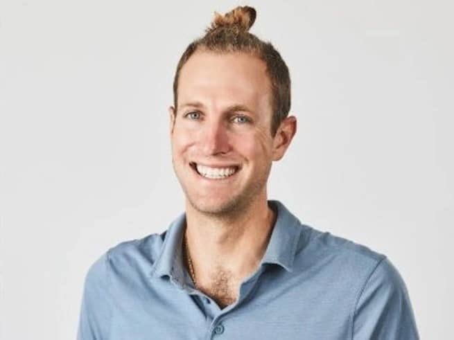 A portrait photo of a smiling man with blonde hair styled in a top knot, wearing a light blue polo shirt against a white background.