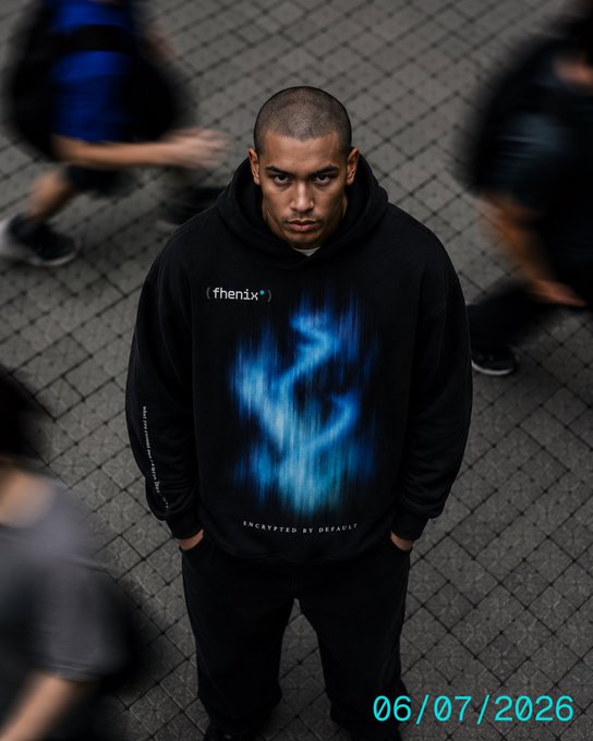 A man wearing a black "fhenix" branded hoodie with blue flame graphics standing on a brick plaza, dated "06/07/2026."