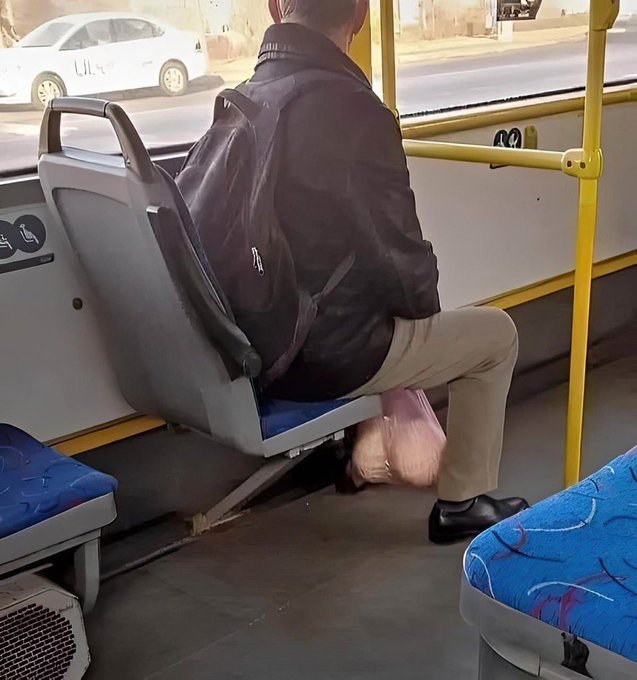 A man in a dark jacket and backpack sits on a public bus, holding a pink plastic bag between his knees—appearing to clutch it protectively like a crypto investor gripping their portfolio. The humor plays on the stereotype of crypto enthusiasts nervously safeguarding their holdings, treating a humble grocery bag with the same intensity as valuable digital assets.