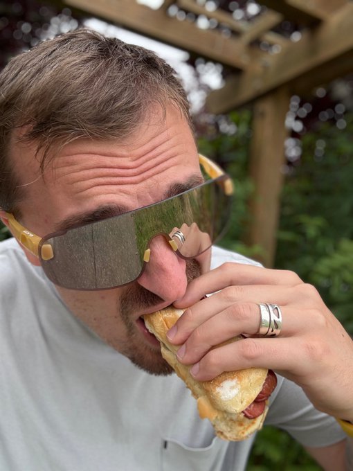 The image features a man wearing sunglasses eating a hot dog while standing outside. The caption reads "Before burgermogging existed, the creator of the @base chain glizzymogged the entire world. Never forget this, Base was first." This message is related to the man's activity and the post about Base being the first person to mog the world with his hot dog.

The image also includes a sandwich in front of him, which he is eating while wearing sunglasses. The background shows trees, indicating that they are outdoors.