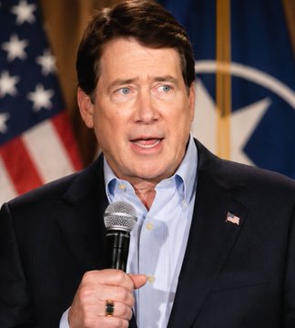 The image shows U.S. Senator Bill Hagerty, a middle-aged man with dark hair wearing a dark blazer over a light blue collared shirt and an American flag lapel pin, speaking into a microphone with an American flag and seal visible in the background. The photo accompanies his statement urging passage of the Clarity Act, where he questions whether Democrats will prioritize mid-term politics over achieving legislative clarity.