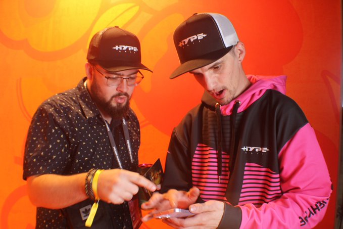 Two men wearing 'HYP3' branded hats and apparel looking down at a phone, set against an orange background.