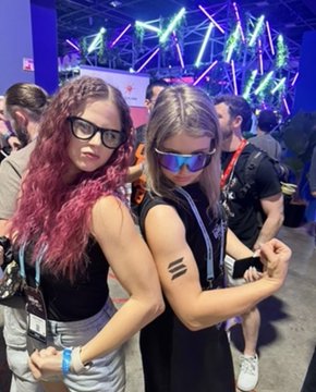 Two friends pose flexing at what appears to be a lively conference or event venue, with vibrant stage lights in the background. The caption reflects on how crypto has connected them with amazing friends worldwide, traveling together from reality shows in Thailand to the WSOP in Vegas to create content.