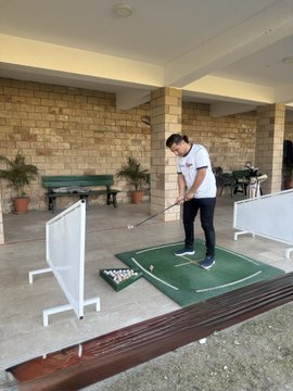 A young man practices his golf swing on a putting green set up under a covered patio area, with a basket of golf balls beside him. The image serves as a lighthearted visual metaphor contrasting leisure activities with the serious claim in the post about building a $900,000 crypto portfolio over 10 years.