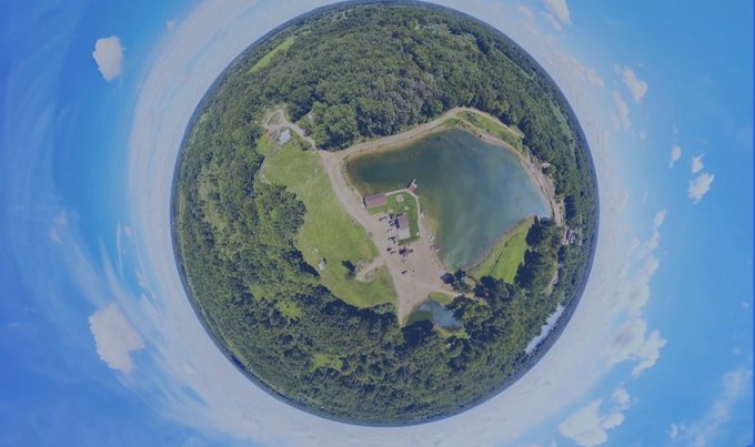 A 360-degree panoramic aerial view presented as a small planet shows a forested area with a lake, a small building, and grassy clearings, surrounded by blue sky and scattered clouds.