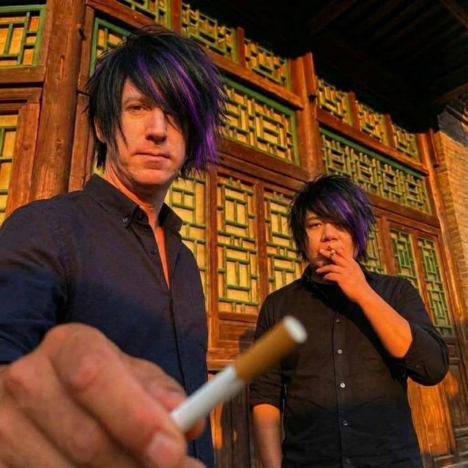 Two men with similar dark and purple-streaked hairstyles, one holding a cigarette, standing in front of an ornate wooden background.