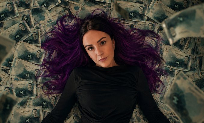 A woman with vibrant purple hair lies on a bed of banknotes, gazing directly at the viewer. The image visually reinforces the post's pitch for a high-yield cashback trading platform ($HYPE), where stacking money symbolizes the lucrative 12% rewards and referral earnings being promoted.