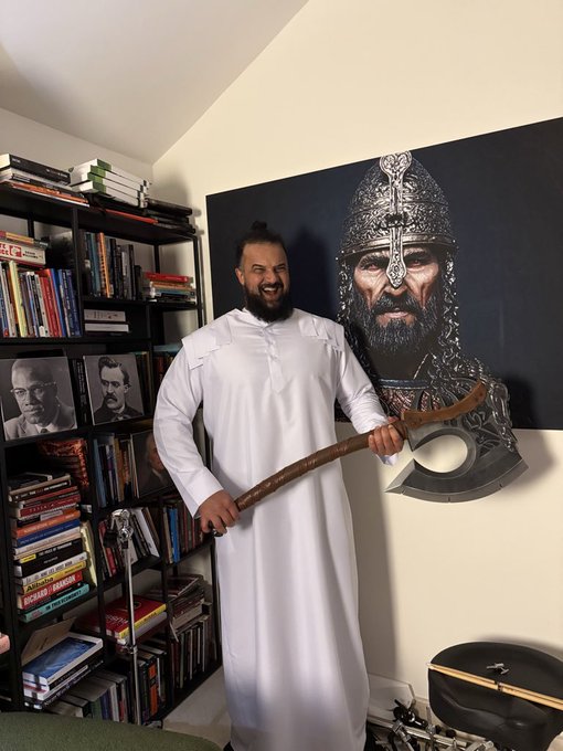 A bearded man in a white robe poses with a large prop axe in front of a bookshelf and a portrait painting of a warrior in chainmail armor.