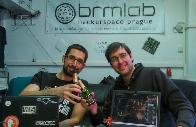 Two men smile while toasting with beer bottles at the brmlab hackerspace in Prague, with a "brmlab hackerspace prague" banner displayed behind them listing the website http://brmlab.cz, Linode #brmlab, and bmlab@bmlab.cz. The image celebrates the 12th anniversary of the first Trezor Bitcoin hardware wallet, which was developed by the Czech-based team at this very hackerspace.