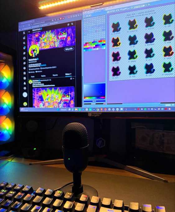 A computer monitor displays digital art software with a colorful image on the left and a sprite sheet of identical cat-like figures on the right, with a microphone and RGB-lit keyboard in the foreground.