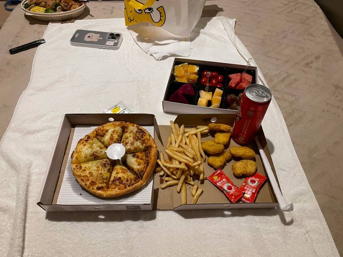 A meal laid out on a white towel, featuring a pizza, french fries, chicken nuggets, a fruit box, and a red soda can, with a phone case in the background.