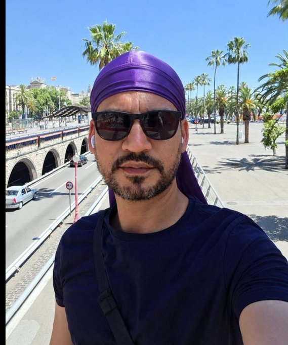 A selfie of a bearded man wearing sunglasses, a purple durag, and earbuds, standing on a sunny day with palm trees and a road with arched tunnels behind him.