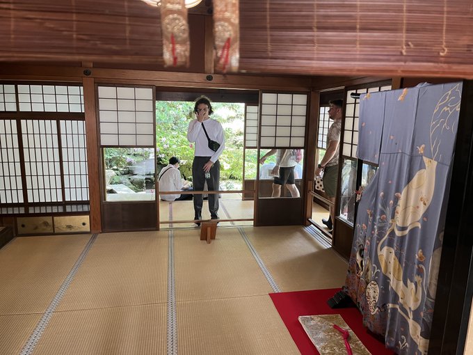 A traditional Japanese-style room with tatami mats, shoji screens, and a decorative curtain, with people visible outside the open doorway in a garden setting.